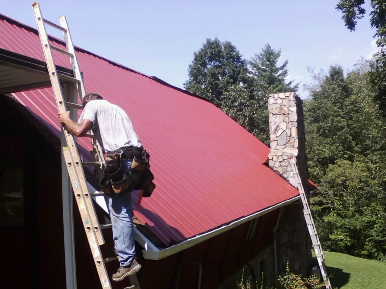 Roofer on a ladder beside a red metal roof and stone chimney — Soffit & Fascia Repair by Roofing Brothers in New Vernon